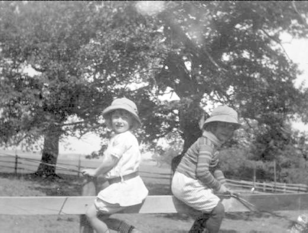 Zwei junge Jungs in Hüten stehen in einem grünen Park und halten Baseballschläger in der Hand, mit Bäumen und einem Zaun im Hintergrund, auf einem Schwarz-Weiß-Foto.