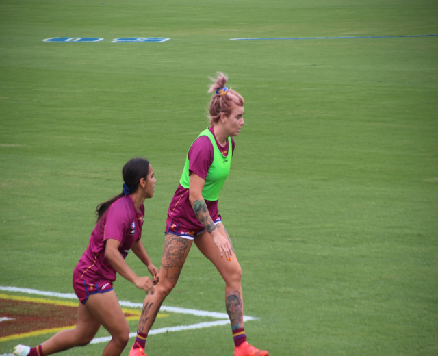 Zwei Frauen in Sportbekleidung spielen Fußball auf einem Feld, eine in einem grünen T-Shirt und Shorts, die andere in einem pinken Kleid, beide mit sichtbaren Tattoos an Händen und Füßen.