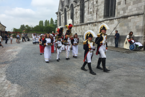 Eine Gruppe uniformierter Personen, darunter einige im Rollstuhl und eine Person mit einem Kinderwagen, marschiert auf einer Straße in Frankreich in einer Militärparade, mit einem Gebäude, Bäumen und einem bewölkten Himmel im Hintergrund.