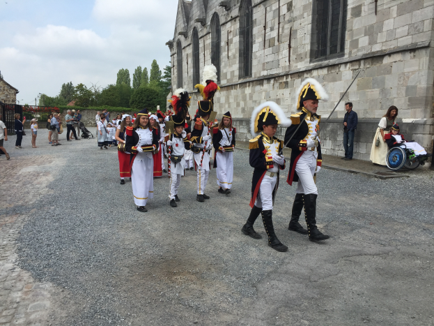 Eine Gruppe uniformierter Personen, darunter einige im Rollstuhl und eine Person mit einem Kinderwagen, marschiert auf einer Straße in Frankreich in einer Militärparade, mit einem Gebäude, Bäumen und einem bewölkten Himmel im Hintergrund.