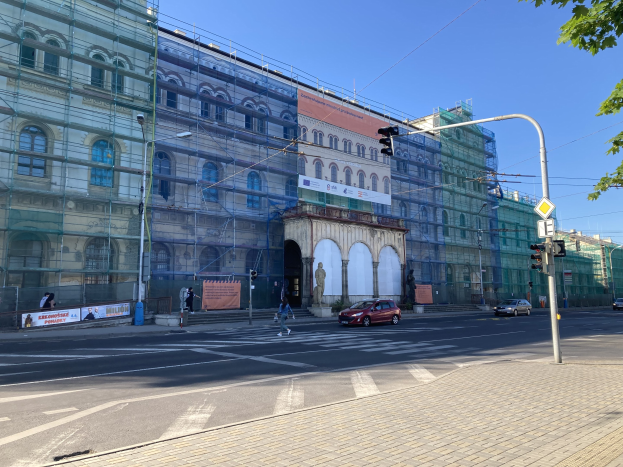 Großes Gebäude mit Gerüst während der Renovierung umgeben von Fahrzeugen, Passanten, Ampeln, Hinweisschildern, Straßeninfrastruktur, Bäumen und einem klaren blauen Himmel.