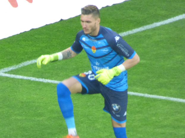 Ein Mann in einem blauen Hemd und grünen Handschuhen steht auf einem Fußballfeld mit einem weißen und roten Banner im Hintergrund, trägt Fußballhandschuhe.