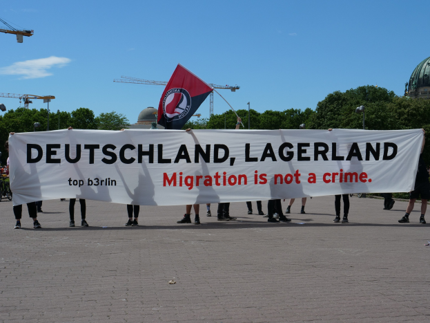 Eine Gruppe von Menschen mit einem Transparent mit der Aufschrift "Deutschland, Lagerland Migration ist kein Verbrechen" und einer Flagge, im Hintergrund sind Laternenpfähle, Laternen, Bäume, Kräne, ein Gebäude und ein bewölktes Himmel zu sehen.