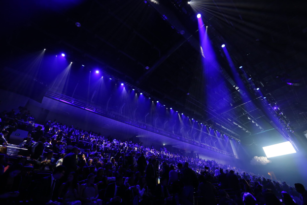 Eine große Menschenmenge vor einer Bühne mit Deckenbeleuchtung, mit einem Bildschirm auf der rechten Seite zu sehen.