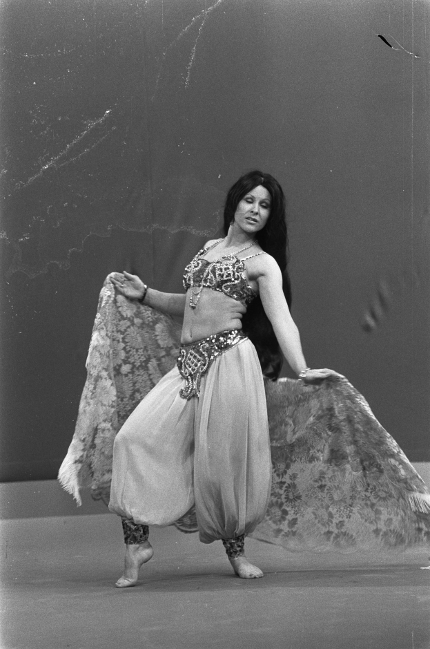 Eine Frau in einem langen, gemusterten Bauchtanzkleid tanzt mit geschlossenen Augen und ausgestreckten Armen vor einer Wand.