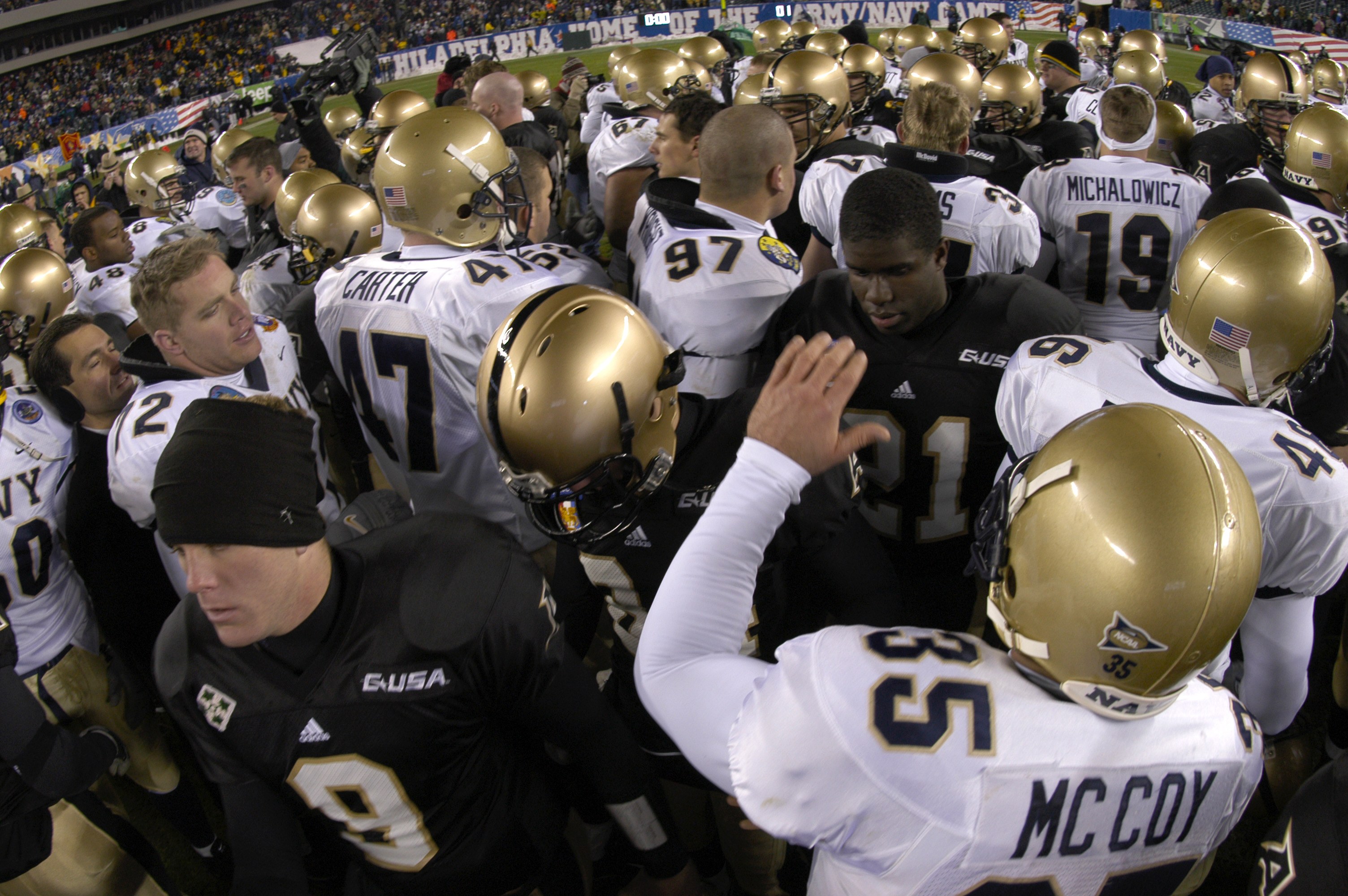 Fußballspieler in Helmen auf einem Rasenfeld versammelt mit Zuschauern auf den Rängen und Bannern im Hintergrund.