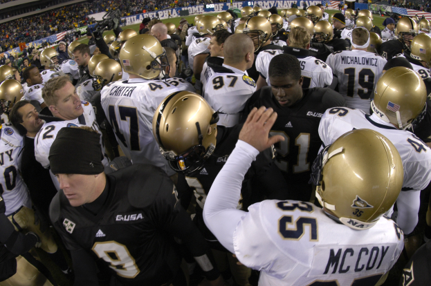 Fußballspieler in Helmen auf einem Rasenfeld versammelt mit Zuschauern auf den Rängen und Bannern im Hintergrund.