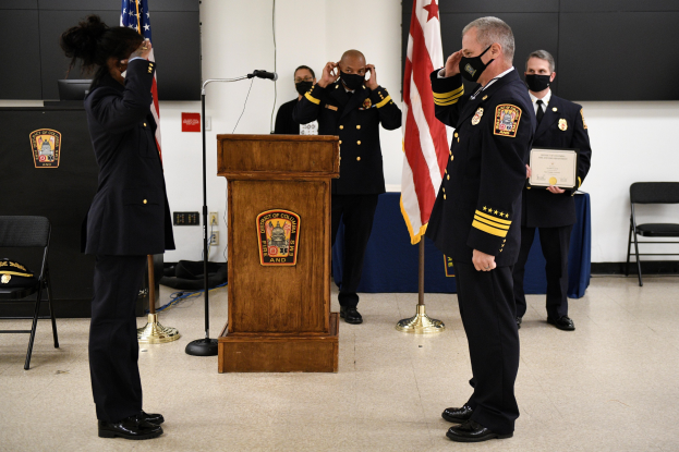 Gruppe uniformierter Personen in Masken vor einem Podium mit einem Mikrofon, umgeben von Fahnen und Möbeln, bei einer Vereidigung eines neuen Feuerwehrchefs.