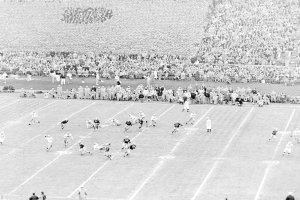 Schwarzes und weißes Foto eines Fußballspiels im Gange, mit Spielern auf dem Feld und einer Zuschauermenge im Hintergrund.