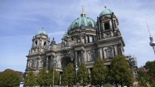 Großes, prunkvolles Berliner Dom-Gebäude in Berlin, Deutschland, mit architektonischen Details wie Fenstern, Säulen, Bögen und Statuen, vor einem bewölkten Himmel, mit Passanten, Bäumen und einem Turm im Vordergrund.