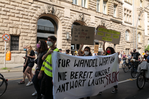 Eine Gruppe von Menschen marschiert auf einer Stadtstraße, einige halten Schilder und andere fahren Fahrräder, im Hintergrund ein historisches Gebäude mitbogigen Fenstern, Säulen und Skulpturen.