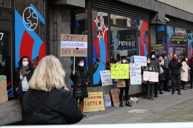Gruppe von Menschen mit Masken, die Schilder und Plakate vor einem Gebäude mit Glasfront halten, mit Kameras und einem Hund, der gegen eine deutsche Regierungspolitik protestiert.