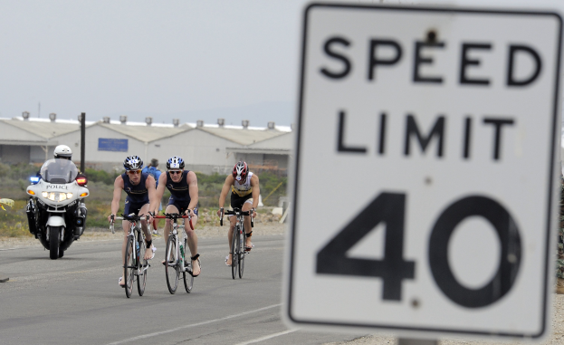 Gruppe von Radfahrern in Helmen, die auf einer Straße mit einem Geschwindigkeitsbegrenzungsschild fahren, mit Gebäuden und Bäumen im Hintergrund unter einem klaren blauen Himmel.
