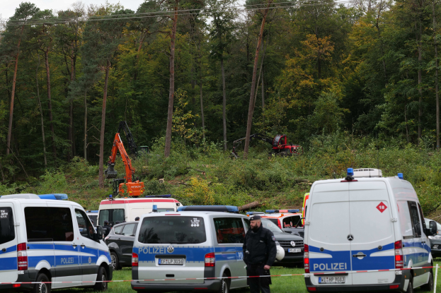 Eine Gruppe von Polizeiwagen auf einem Parkplatz mit einer Person in der Nähe, Bäume und eine Baggerschaufel im Hintergrund unter einem sichtbaren Himmel.