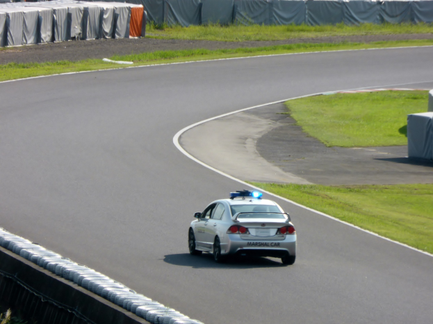 Polizeiauto fährt auf einer Rennstrecke mit Gras auf beiden Seiten, mit Menschen, Zelten und Gegenständen im Hintergrund.