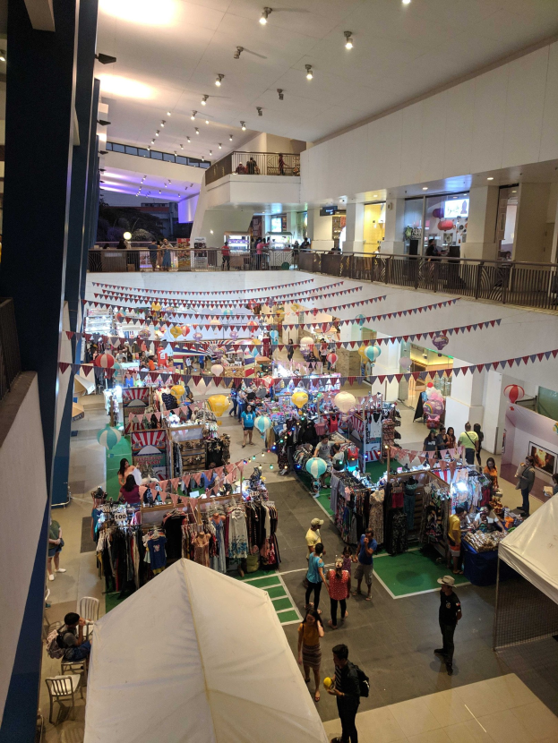 Ein volles Einkaufszentrum mit Ständen, hängenden Kleidern, Dekorationsfahnen, Ballons, sitzenden und stehenden Menschen, Geländern, Säulen und Deckenlampen, das an einen Weihnachtsmarkt im Mall of Asia erinnert.