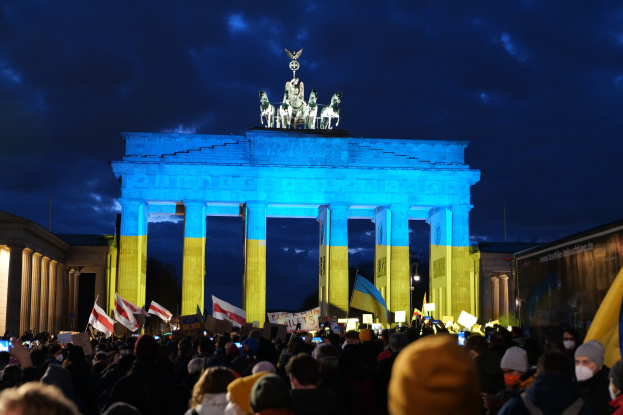 Menge mit Flaggen und Plakaten vor dem Brandenburger Tor, mit einem Banner auf der rechten Seite mit protestrelevanten Text.