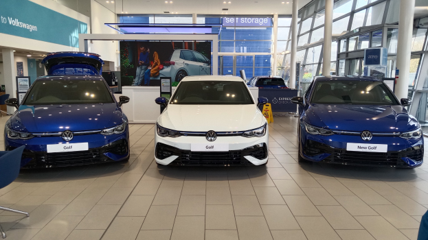 Volkswagen-Ausstellung mit drei blauen und weißen Autos, einem Stuhl und einem Tisch mit Gegenstaenden auf der linken Seite und Informationsplaenen mit Bildern im Hintergrund.