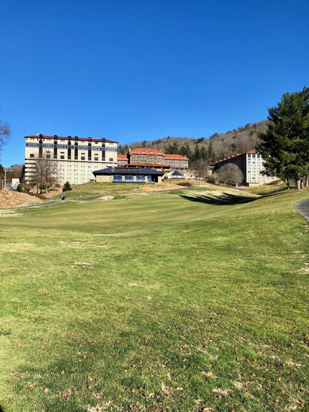 Golfplatz mit einem Resort-Hotel im Hintergrund, umgeben von grünem Gras, Bäumen und einer Straße unter einem blauen Himmel.
