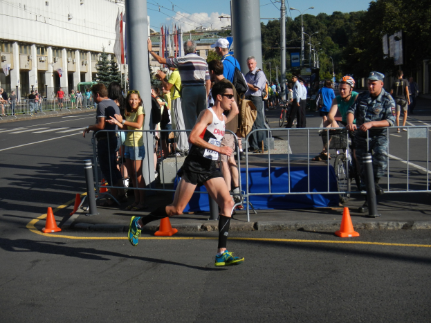 Ein Mann läuft bei einem Marathon auf einer Stadtstraße, umgeben von Zuschauern mit Fahrrädern, Verkehrspfosten, Absperrungen, Flaggen, Straßenlaternen, Strommasten, Bäumen, Gebäuden und einem bewölkten Himmel.