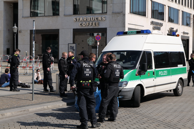Eine Gruppe von Polizisten steht vor einem Starbucks-Café mit einem Van auf der rechten Seite und einigen Menschen auf der linken Seite; im Hintergrund sind ein Gebäude, ein Schild, ein Laternenmast und ein Zaun zu sehen.