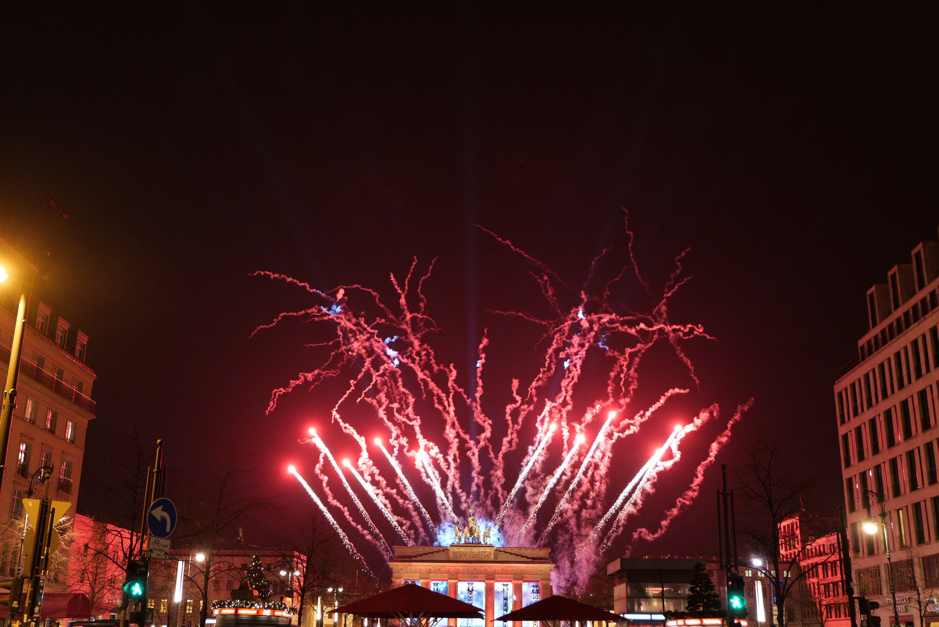 Eine belebte Stadtstraße während der Silvesterfeierlichkeiten in Berlin, mit Menschenmassen, Fahrzeugen, beleuchteten Gebäuden, Feuerwerk am Himmel und festlicher Dekoration.