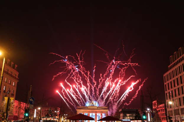 Eine belebte Stadtstraße während der Silvesterfeierlichkeiten in Berlin, mit Menschenmassen, Fahrzeugen, beleuchteten Gebäuden, Feuerwerk am Himmel und festlicher Dekoration.