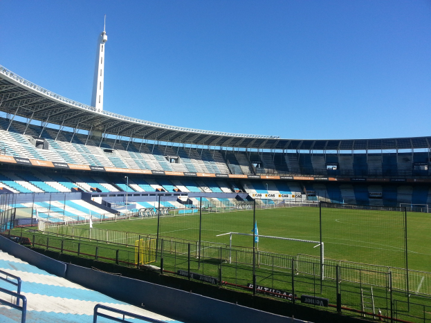 Großes Stadion mit einem von einem Zaun umgebenen Fußballfeld, einem Turm im Hintergrund und einem klaren blauen Himmel darüber.