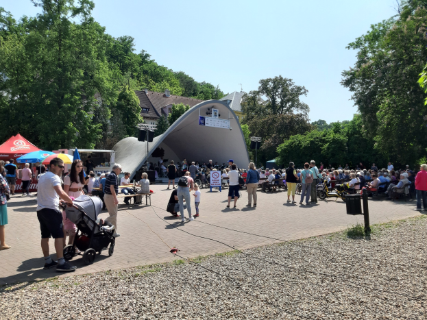 Eine Menschenmenge steht vor einem Gebäude bei der Fest-Eröffnung, einige sitzen auf Bänken, andere stehen; ein Kinderwagen ist im Vordergrund zu sehen, sowie Bäume, Gebäude und einen klaren blauen Himmel im Hintergrund, sowie Zelte links und einen Mülleimer rechts.
