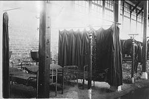 Ein Schwarz-Weiß-Foto eines Krankenhauszimmers mit mehreren Betten, Stangen, Vorhängen und einer Wand im Hintergrund, mit Text unten.