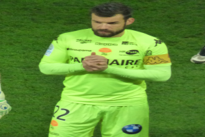 Ein Mann in einem gelben Torwarttrikot, der eine grüne T-Shirt und Shorts trägt, steht auf einem Fußballfeld und klatscht in die Hände, wobei ein weiteres Bein auf der linken Seite des Bildes zu sehen ist.