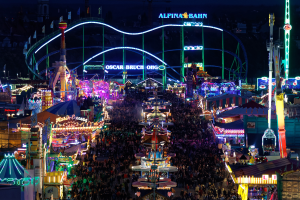 Eine große Menschenmenge bei einem nächtlichen Karneval, beleuchtet von bunten Lichtern, umgeben von Vergnügungsgeräten, Pfählen, Fahnen und Namensschildern, mit einem dunklen Hintergrund.