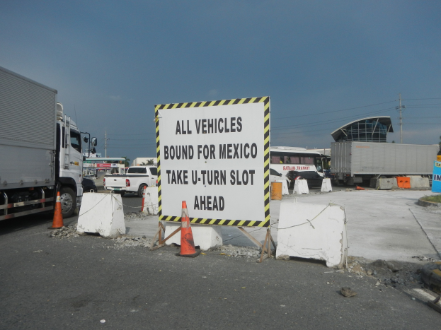 Ein Lkw auf einem Parkplatz neben einem Schild mit der Aufschrift "Alle Fahrzeuge nach Mexiko", mit verstreuten Konen, Brettern und anderen Gegenständen und im Hintergrund Gebäude, Bäume und Masten unter einem klaren blauen Himmel.