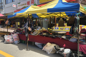 Ein Straßenmarkt mit Zelten, die verschiedene Gegenstände wie Taschen und Hüte präsentieren, vor einem Hintergrund aus Gebäuden mit Fenstern und Schildern.