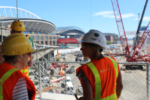 Zwei Frauen in Sicherheitswesten, Helmen, Schutzbrillen und Schutzhelmen stehen auf einer Baustelle, eine hält ein Objekt, mit Maschendrahtzaun dahinter und Fahrzeugen, Kränen, Gebäuden, Bäumen und einem bewölkten Himmel im Hintergrund.