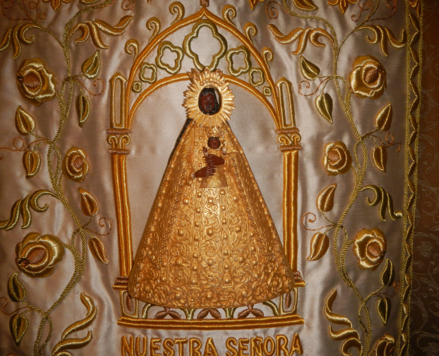 Goldbrokate Chasuble mit einer serenen Frau in einem hochgeschlossenen, langärmligen Kleid mit einem Dutt, umgeben von detaillierten Designs, Text und goldenen Bordüren.