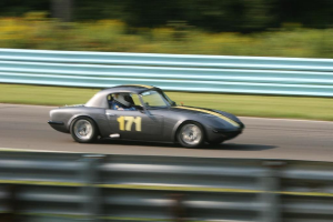Ein Auto mit einem Helm tragenden Fahrer fährt eine Straße entlang, die von Barrieren gesäumt ist und Bäume im Hintergrund hat.