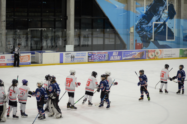 Group of young people playing ice hockey on an indoor rink, wearing helmets and holding sticks, with a person standing on the left and banners, pillars, and glass walls in the background.