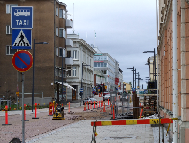 Stadtstraße mit Gebäuden, Straßenlaternen, Verkehrskegeln, Fahrzeugen, Absperrpoller, Bäumen und einer Baustelle mit Verkehrsschildern unter einem bewölkten Himmel.