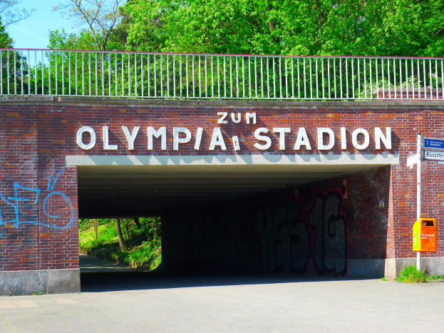 Der Eingang zum Olympiastadion in Berlin, Deutschland, mit einer Brücke mit Text, einem Metallzaun, einer Tafel, einem Kasten, Pflanzen, Gras, einer Baumgruppe und einem bewölkten Himmel.