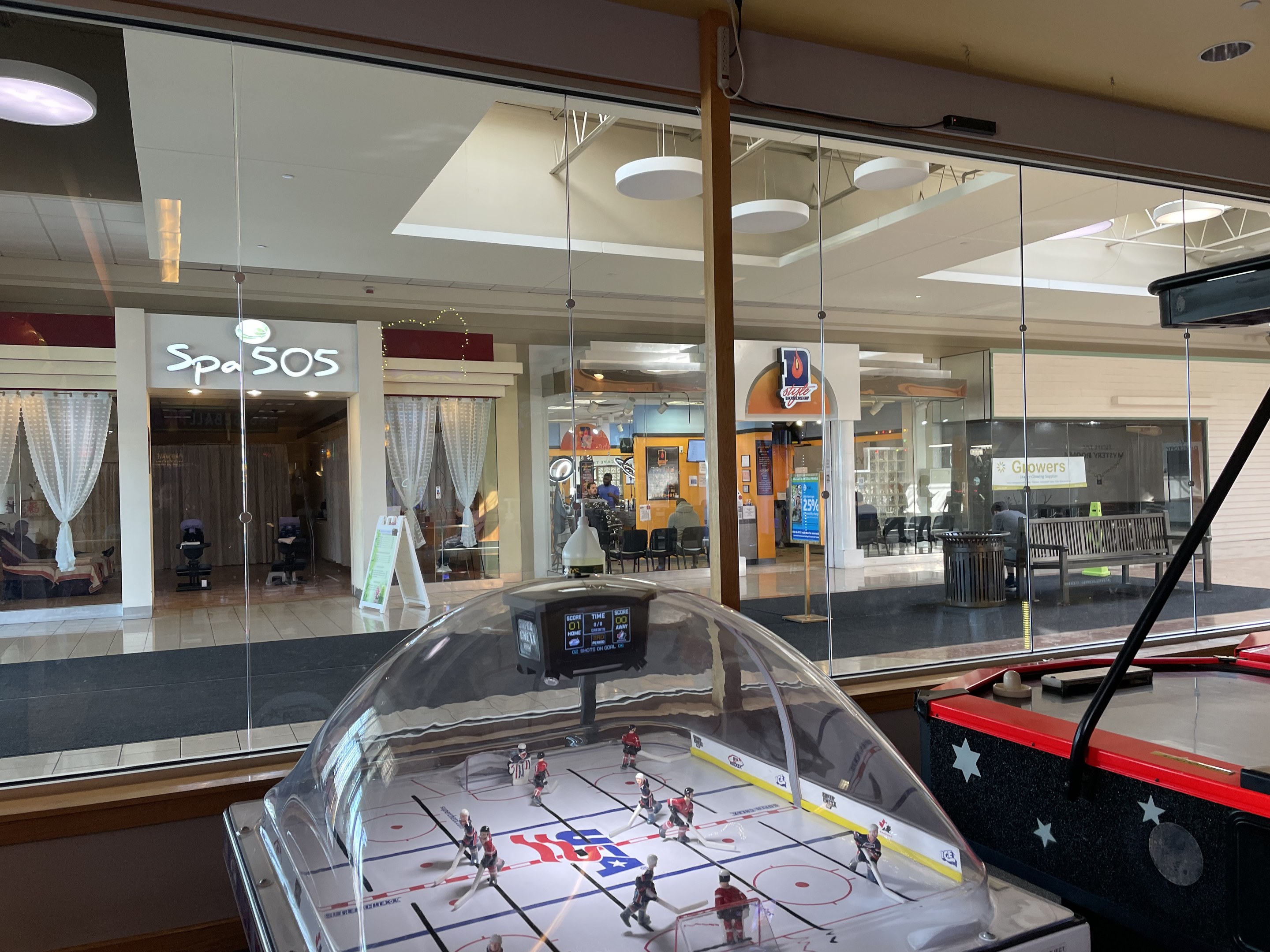 Eine Eishalle in einem Einkaufszentrum, umgeben von Glaswänden mit Menschen, Möbeln und Beschriftung draußen und einem Air-Hockey-Tisch auf der rechten Seite.