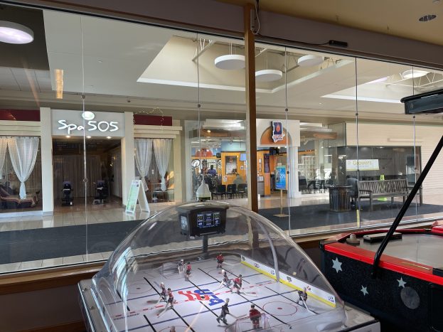 Eine Eishalle in einem Einkaufszentrum, umgeben von Glaswänden mit Menschen, Möbeln und Beschriftung draußen und einem Air-Hockey-Tisch auf der rechten Seite.