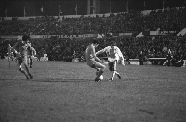 Schwarzes und weißes Bild von Männern, die Fußball in einem Stadion mit Zuschauern und Fahnen im Hintergrund spielen.