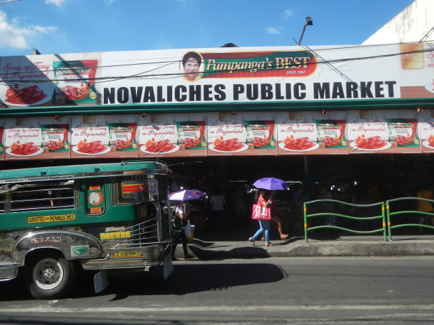 Ein Bus fährt an einem Gebäude mit der Aufschrift "Novaliches Public Market" vorbei, mit Passanten, Schirmen, einem Geländer und städtischen Elementen im Hintergrund.