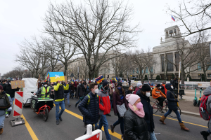 Eine große Gruppe von Menschen nimmt an einer Protestdemo auf einer Straße in Washington, D.C. teil, wobei einige Schilder und Plakate halten und andere Fahrräder fahren, sowie Schilder, Bäume und ein klarer blauer Himmel im Hintergrund zu sehen sind.
