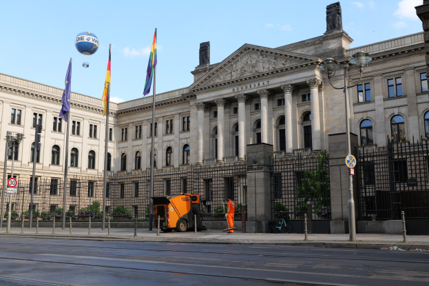 Großes modernes Gebäude mit vielen Fenstern, der Bundestag in Berlin, umgeben von Straßeninfrastruktur, Füßgängern, Flaggen und Bäumen unter einem bewölkten Himmel.