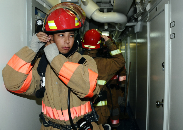 Gruppe von Feuerwehrleuten in Uniform, die zusammen in einem Raum mit einer Tür auf der rechten Seite und sichtbaren Rohren im Hintergrund stehen, wahrscheinlich während eines Trainings.