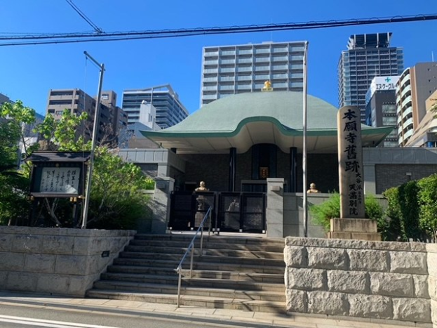 Großes grünoffenes Gebäude in städtischer Umgebung mit umgebenden Treppen, Geländern, Beschilderung, Grünflächen und einer Straße im Vordergrund, Wolkenkratzer im Hintergrund unter einem klaren Himmel.