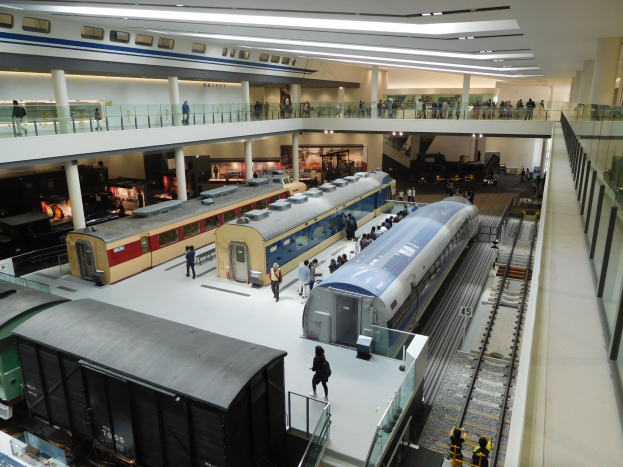 Eine Museumsausstellung, die verschiedene Züge in unterschiedlichen Größen und Farben zeigt, mit Menschen, die von Boden, Geländern und Säulen aus beobachten.