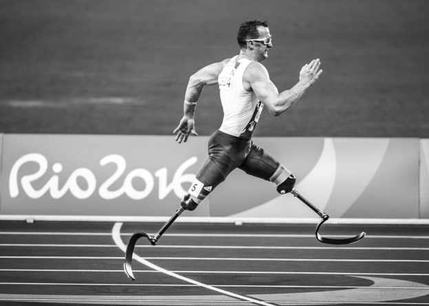 Schwarz-weiß-Foto eines Mannes mit Prothese, der auf einer Laufbahn läuft, mit einer Tafel im Hintergrund, auf der "Rio 2016 Paralympische Spiele" steht.